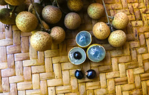 Kahverengi arka planda izole edilmiş bambu sepetinde yaprakları olan taze yerel longan meyvesi ve soyulmuş longan. Asya tropikal meyvesi. Bahçedeki Longan hasadı