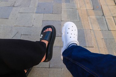 Otururken iki farklı ayakkabı pozisyonu. Beyaz ayakkabılar ve siyah sandaletler.