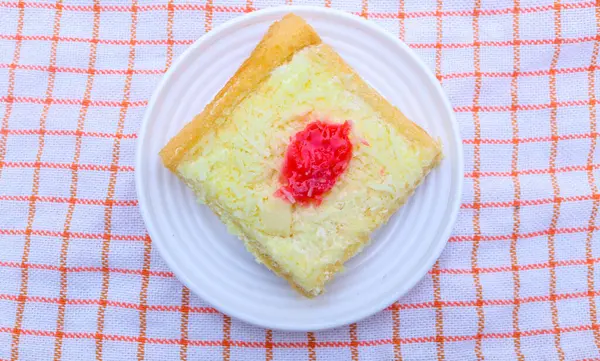 Üzerinde çilek reçeli ve rendelenmiş peynir olan bir dilim kare cheesecake beyaz tabak ve peçeteyle servis edilir. Ahşap masa arkaplanında izole