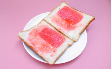 Bir tabakta çilek reçeli ve pembe zemin üzerinde iki dilimlenmiş ekmek ile beyaz ekmek kaplayın. Basit kahvaltı. Reçelli tost..