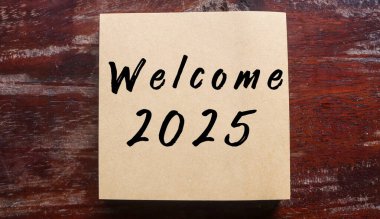 Tahta masada 2025 numaralı hoş geldin yazılı kahverengi yapışkan not. Yeni yıl 2025 kavramını hoş geldiniz.