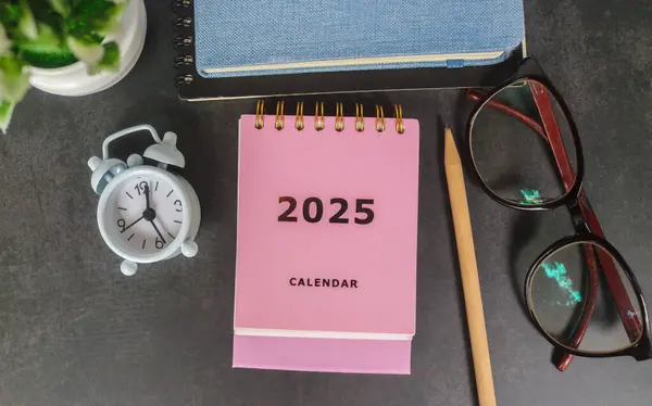 Pembe takvim 2025, ofis masasında defter, gözlük, kalem ve çalar saatle. Yeni yıl 2025 ofis masası konseptiyle