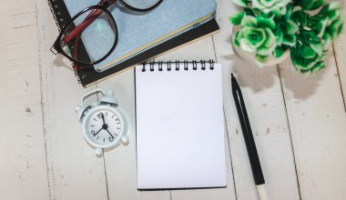 Beyaz ofis masasında, içinde kitaplar, kalem, bitki, gözlük, saat ve diğer şeylerin yazılı olduğu boş defter. Metni ortaya koymak için boş defter sayfası. Üst manzara, düz uzanma.