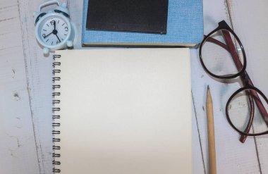 Beyaz ofis masasında, içinde kitaplar, kalem, bitki, gözlük, saat ve diğer şeylerin yazılı olduğu boş defter. Metni ortaya koymak için boş defter sayfası. Üst manzara, düz uzanma.