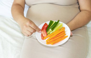 Ellerinde avokado çöreği bebek kıyafetleri olan ya da koluna krem sürerek koluna krem sürmüş hamile bir kadın.