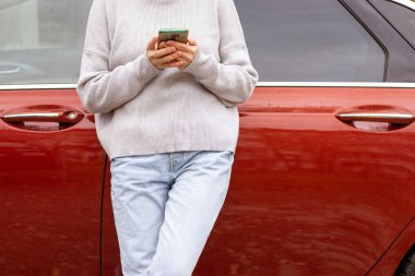 woman in front of lateral door red car in warm pullover blue jeans holding hands on waist,typing message smartphone,one leg raised up,sole of boot open door searching in vehicle interior back view.