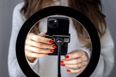 woman girl young blogger vlogger talking with followers hand holding phone in middle of selfie light round lamp.blonde hair red nails female explaining moving hands from side to side or typing 