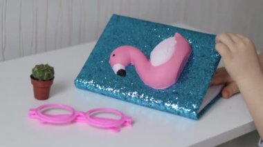 Masanın üzerinde parıltılı ve yumuşak oyuncak pembe flamindo, minimalist beyaz masa, küçük kaktüs bitkisi ve pembe gözlüklü mavi defter. Çocuk çocuk ya da kız Teeneager el yumuşak flamindo açık not defterine dokunuyor.