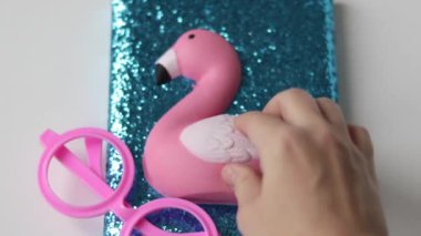 Masanın üzerinde parıltılı ve yumuşak oyuncak pembe flamindo, minimalist beyaz masa, küçük kaktüs bitkisi ve pembe gözlüklü mavi defter. Çocuk çocuk ya da kız Teeneager el yumuşak flamindo açık not defterine dokunuyor.