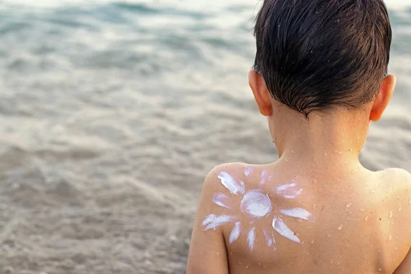 kid boy preschooler with drawn sun shape from cream lotion on shoulder against sea ocean tan red skin hand up family vacation summer time.sun block protection white cream uv rays safe for skin care