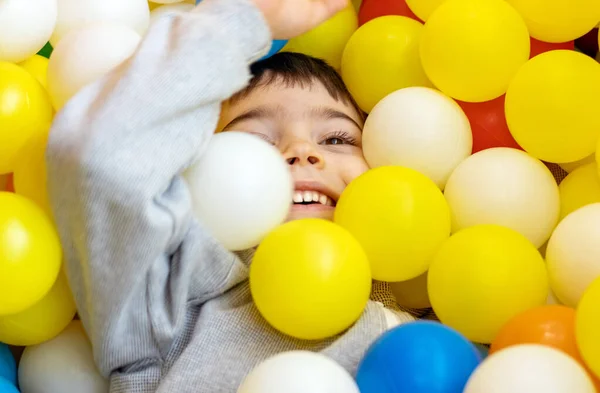 kid having fun in play center pool with colorful many balls.happy smiling child preschooler boy throwing tossing up plastic balls playground interior inside mall.soft ocean plastic balls cover face