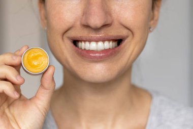 Dudak kremi süren ya da doğal malzemelerden sürtünen bir kadın. Yüzünün yarısı öpücükle gülümseyen bir jest. Küçük kavanozlardaki kızın dudak kremi ve fırçası.