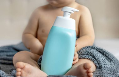 Bebek şampuan banyosu jeli şişesi yumuşak oyuncak ayı reklam afişi reklamları. Bebek bebek yatakta oyun oynuyor. Portre çocuğu elleriyle kafa boşluğunda komik yüz ifadeleri yapıyor.