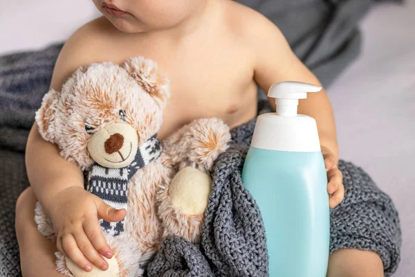 Bebek şampuan banyosu jeli şişesi yumuşak oyuncak ayı reklam afişi reklamları. Bebek bebek yatakta oyun oynuyor. Portre çocuğu elleriyle kafa boşluğunda komik yüz ifadeleri yapıyor.