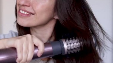 Gülümseyen genç bir kadın modern bir saç fırçası kullanıyor. Saç kurutma makinesi ve hacimsel dönüşümlü kafa yavaş çekimde. Kurutma makinesinin termal ve seramik kaplaması. Kaküllerden önce karışık saç.