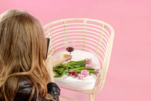 Çekici bir kadın pembe arka plandaki şarapla bardak alıyor. Siyah deri ceketli, sandalyede oturan ve kompozisyon fotoğrafı çeken bir kız.