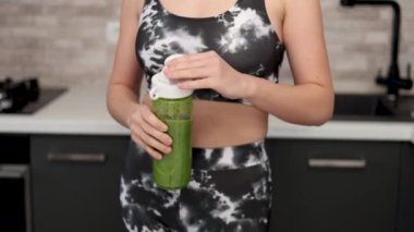 Kadın mutfakta kereviz, elma, mandalina, kivi ve ıspanak karışımından sağlıklı bir içecek hazırlıyor. Spor takım elbiseli kız blender kabında meyve, su ve karışım ekliyor. Kadın ezilmiş meyve, ince vücut 4k.