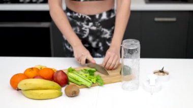 Kadın mutfakta kereviz, elma, mandalina, kivi ve ıspanak karışımından sağlıklı bir içecek hazırlıyor. Spor takım elbiseli kız blender kabında meyve, su ve karışım ekliyor. Kadın ezilmiş meyve, ince vücut 4k.