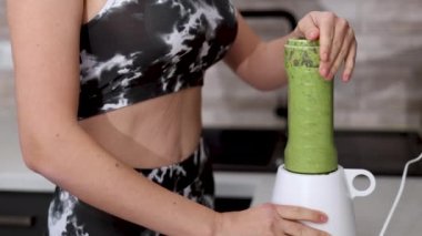 Kadın mutfakta kereviz, elma, mandalina, kivi ve ıspanak karışımından sağlıklı bir içecek hazırlıyor. Spor takım elbiseli kız blender kabında meyve, su ve karışım ekliyor. Kadın ezilmiş meyve, ince vücut 4k.