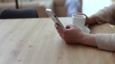 Milenyum kadını mutfakta oturmuş kahve içiyor, akıllı telefon kullanıyor, gülümsüyor. Modern ev içi, bebek bebek bebek arka planda, anneyi rahatlatıyor. El şekerini kaşıkla karıştırıyor.