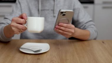 Milenyum kadını mutfakta oturmuş kahve içiyor, akıllı telefon kullanıyor, gülümsüyor. Modern ev içi, bebek bebek bebek arka planda, anneyi rahatlatıyor. El şekerini kaşıkla karıştırıyor.
