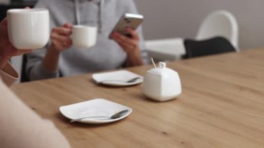Milenyum kadını mutfakta oturmuş kahve içiyor, akıllı telefon kullanıyor, gülümsüyor. Modern ev içi, bebek bebek bebek arka planda, anneyi rahatlatıyor. El şekerini kaşıkla karıştırıyor.