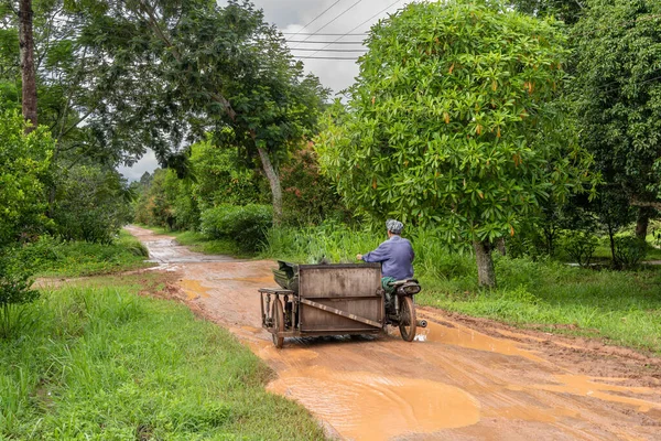 Behind a rural man driving a motorcycle along a dirt road drenched in rain.