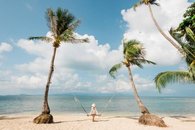 A woman in a white hat sits on a swing among the palm trees on Leela Beach on Koh Phangan Island and watch the sea