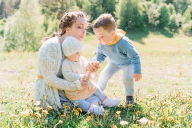 A young mother sits on the grass with her three-year-old son and one-year-old daughter, they blow on dandelions
