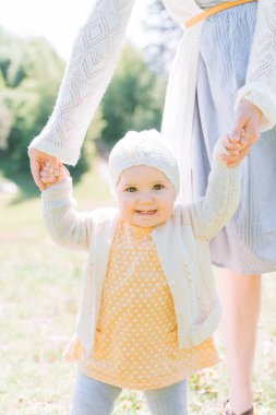 close-up portrait of a baby girl in knitted clothes holding on to her mother's hands and standing on a green meadow on a bright sunny day