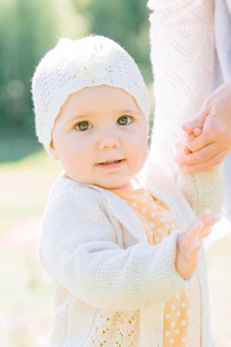 close-up portrait of a baby girl in knitted clothes holding on to her mother's hands and standing on a green meadow on a bright sunny day