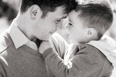 Üç yaşında oğlu olan genç bir babaya yakın çekim, açık havada sarılıp gülümsüyor, siyah-beyaz. Mutlu bir çocuk yetiştirme konsepti.