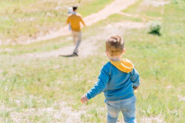 A boy of three years old follows his father carrying his youngest daughter in his arms along a green hilly lawn on a sunny day, view from the back
