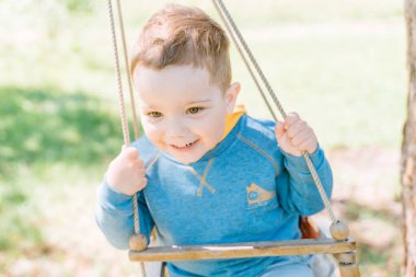 Sarı mavi elbiseli mutlu üç yaşındaki çocuk kırsalda güneşli bir günde salıncağa sımsıkı tutunuyor. Kapatın. Çocukların güveni, kararlılığı, güvenliği.