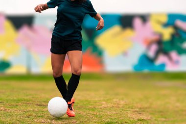 Futbol topunu tekmelemek için koşan esmer kadın futbolcunun bacakları görünüyor. Açık havada yeşil bir çimenlikte, renkli bir arka planda. Spor kıyafetleriyle. Spor ve sağlık reklamı