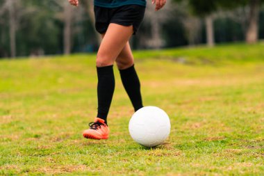 Futbol topunu tekmelemek için koşan esmer kadın futbolcunun bacakları görünüyor. Yeşil bir çimenlikte, spor kıyafetleriyle. Spor ve sağlık reklamı.