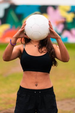 Güzel bir esmer kadın futbolcunun poz verişini, bir futbol topuyla oynayışını ve yüzünü kapamasını görmek. Açık yeşil bir çimenlikte, renkli bir arka planı var. Spor kıyafetleri..