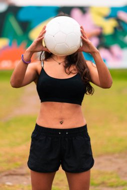 Güzel bir esmer kadın futbolcunun poz verişini, bir futbol topuyla oynayışını ve yüzünü kapamasını görmek. Açık yeşil bir çimenlikte, renkli bir arka planı var. Spor kıyafetleri..