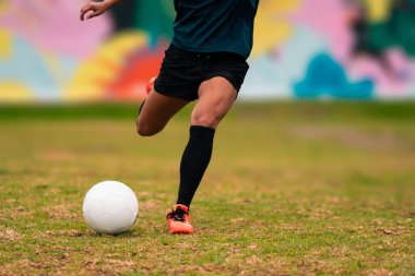 Futbol topunu tekmelemek için koşan esmer kadın futbolcunun bacakları görünüyor. Açık havada yeşil bir çimenlikte, renkli bir arka planda. Spor kıyafetleriyle. Spor ve sağlık reklamı