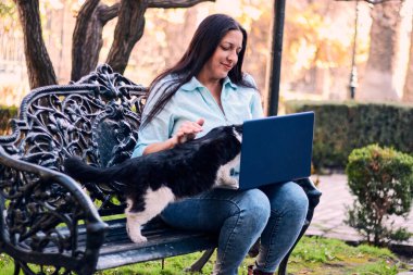 Evinin bahçesinde dizüstü bilgisayarıyla çalışan güzel bir Latin yetişkin kadın kedisi tarafından alay edilip rahatsız ediliyor. tele-çalışma, pazarlama.