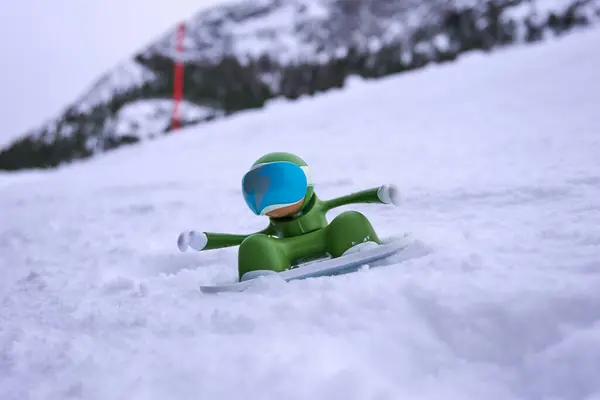Karlı bir dağ sahnesinde çekilmiş mavi gözlüklü, oyuncu, yeşil bir snowboardcu oyuncağı. Kış macerası, yaratıcılık ve çocuksu bir eğlence. Açık hava sporları için mükemmel.