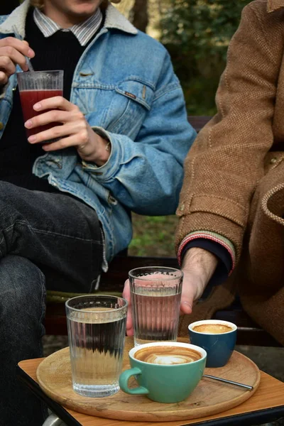 Two white males, one blonde, another  brown haired, drinking coffee and raspberry juice in a public park on a sunny winter day