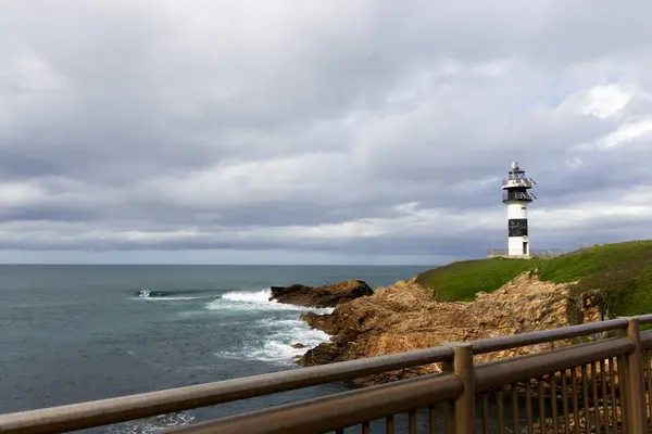 Deniz feneriyle kıyı manzarası bulutlu gökyüzüne karşı kayalık taşar, kıyıda dalgalar kırılır..