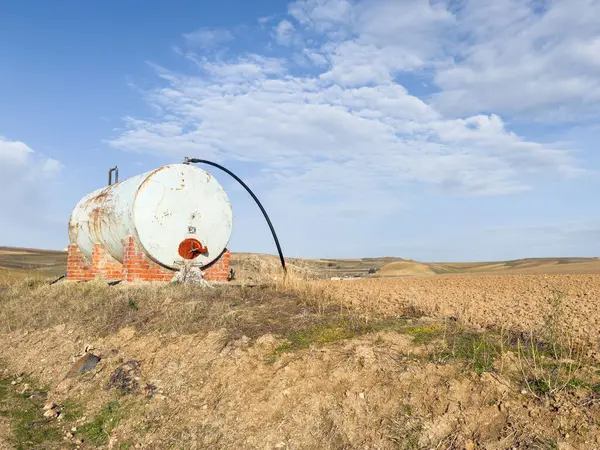 Parçalı bulutlu bir gökyüzünün altında kuru, kahverengi ekinlerin ortasında eski bir yakıt tankını barındıran kırsal bir arazi..