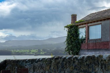 Bu fotoğraf, duvarlarına sarmaşık tırmanan kırmızı tuğlalı tuhaf bir eve açılan eski bir taş duvarı gösteren dingin bir göl kenarı görüntüsü..