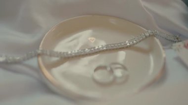 Gelin bileziği ve yüzükler, yanında gelinlik ayakkabıları olan seramik bir tabakta saten üzerinde. Gelinlerin yakın plan fotoğrafları ve düğün davetiyesi..