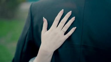 Close-up video shot in slow motion of a brides hands on the back of the groom in a black suit. Bride and groom hugging outside in the day.