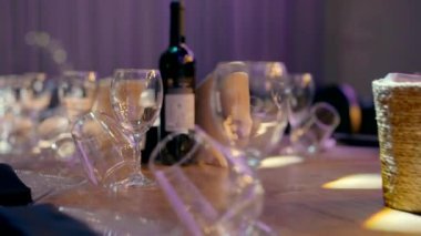 A beautiful wooden dining table for a wedding for many guests. A bottle of wine, wine glasses, plates with black napkins on them on a wooden table in an event hall