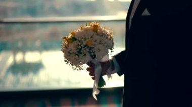 A groom in a suit stands holding a bouquet of white and yellow flowers for the bride on the wedding day.