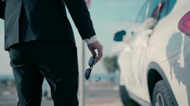 A close-up video shot in slow motion of a groom walking by the white wedding car in the daytime and holding sunglasses.